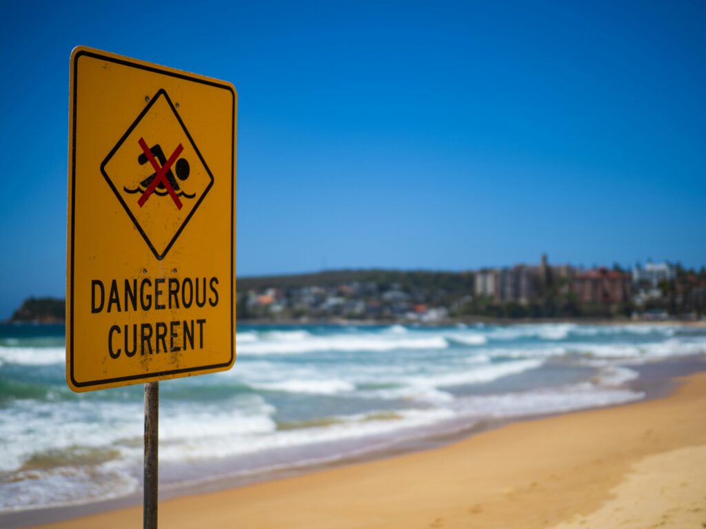 Dangerous current warning sign on Manly Beach in Australia, with blue sky and ocean waves.