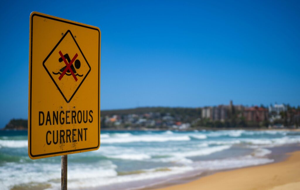 Dangerous current warning sign on Manly Beach in Australia, with blue sky and ocean waves.