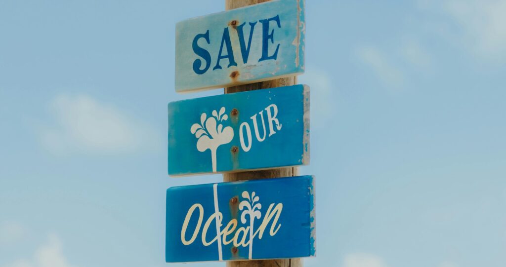 A weathered wooden sign on a beach reads 'Save Our Ocean' against a clear blue sky.
