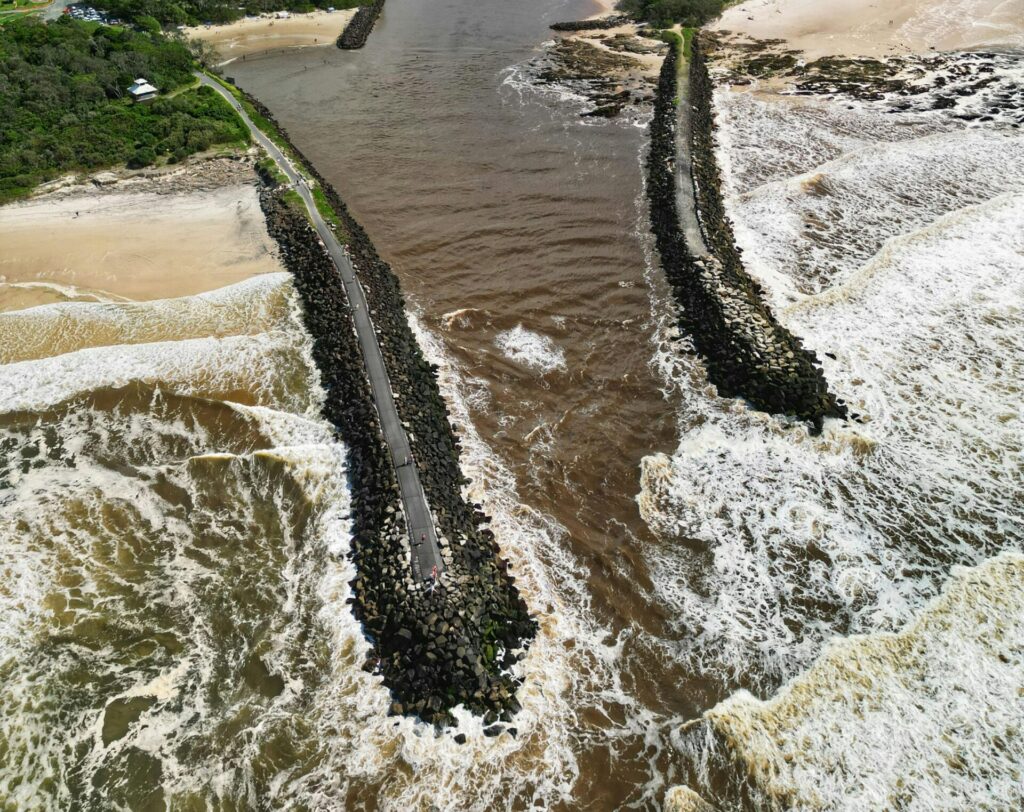 An aerial view of a river estuary meeting the ocean with a visible breakwater structure.