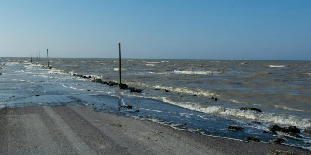 A flooded coastal road surrounded by the sea and horizon under a clear blue sky.