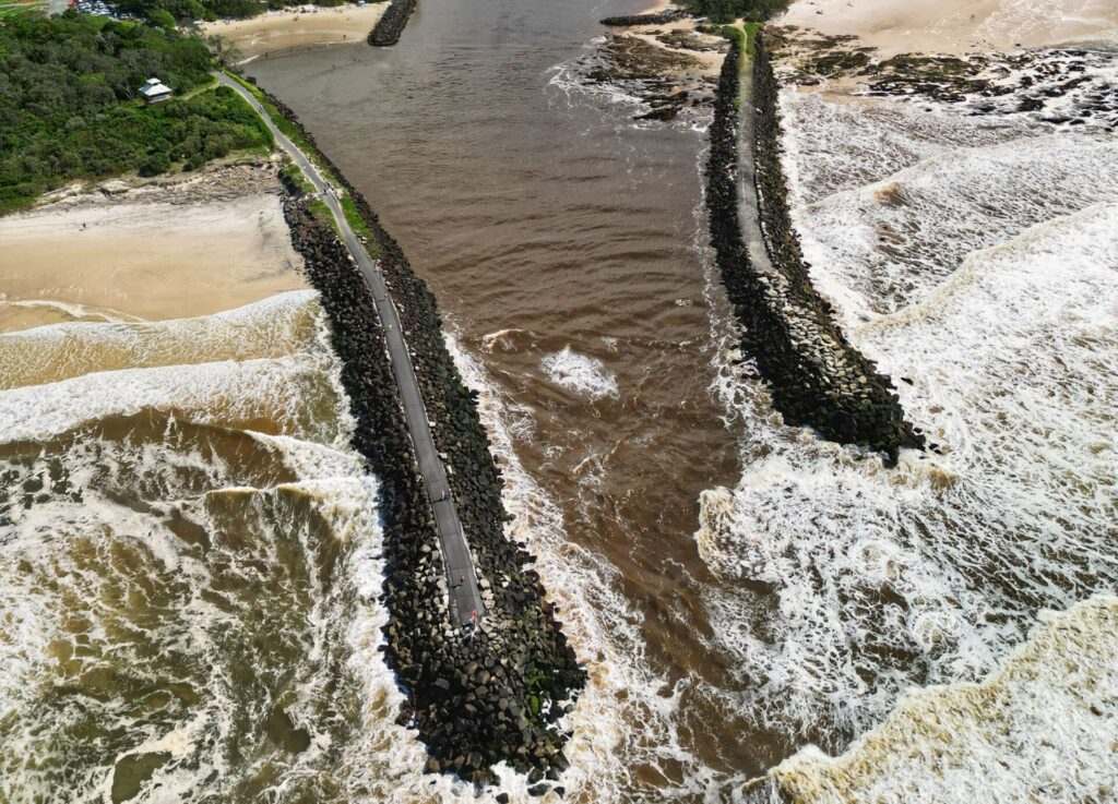 An aerial view of a river estuary meeting the ocean with a visible breakwater structure.