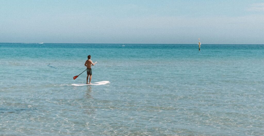 A lone man paddle boarding on a clear blue ocean under a sunny sky, exuding tranquility.